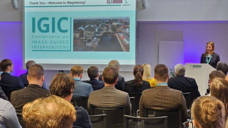 Blick über die Publikumsreihen auf Rednerpult, Rednerin und Präsentation mit Willkommensfolie