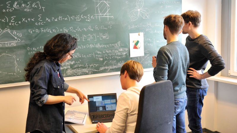 Junge Forschende vor einer Tafel mit mathematischen Formeln. Zwei stehen an der Tafel, zwei arbeiten an einem Laptop davor.