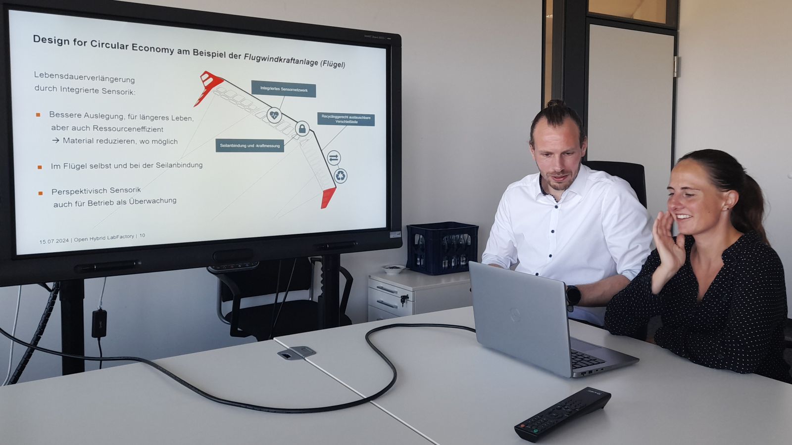 Tim Fröhlich und Nicole Allgaier sitzen am Laptop. Links im Bild zeigt ein Flachbildschirm die Präsentation, auf die beide schauen.