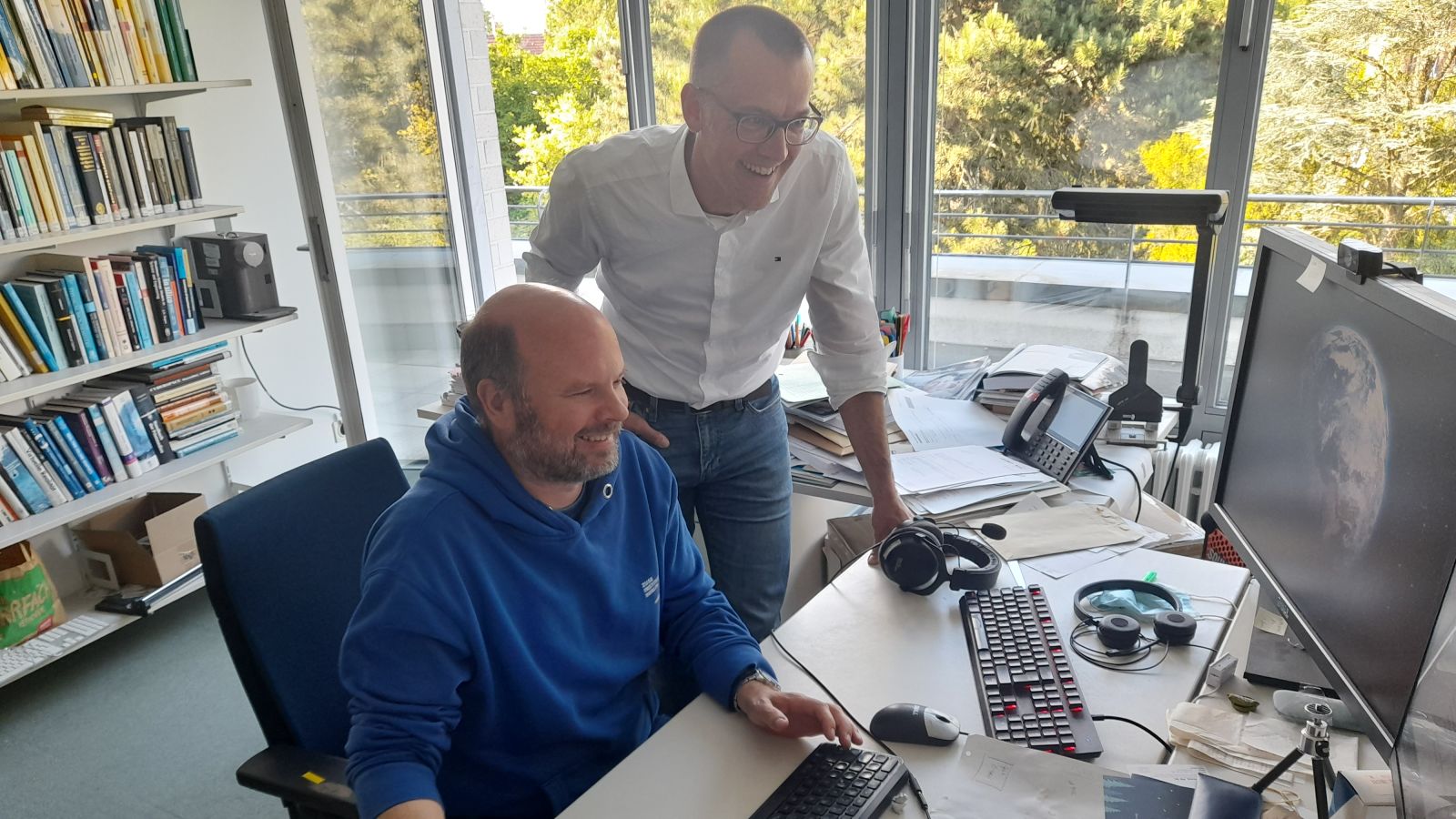 Tim Conrad im blauen Hoodie vor dem Computer sitzend, hinter ihm und über ihn gebeugt steht Hans Lamecker im Weißen Oberhemd.
