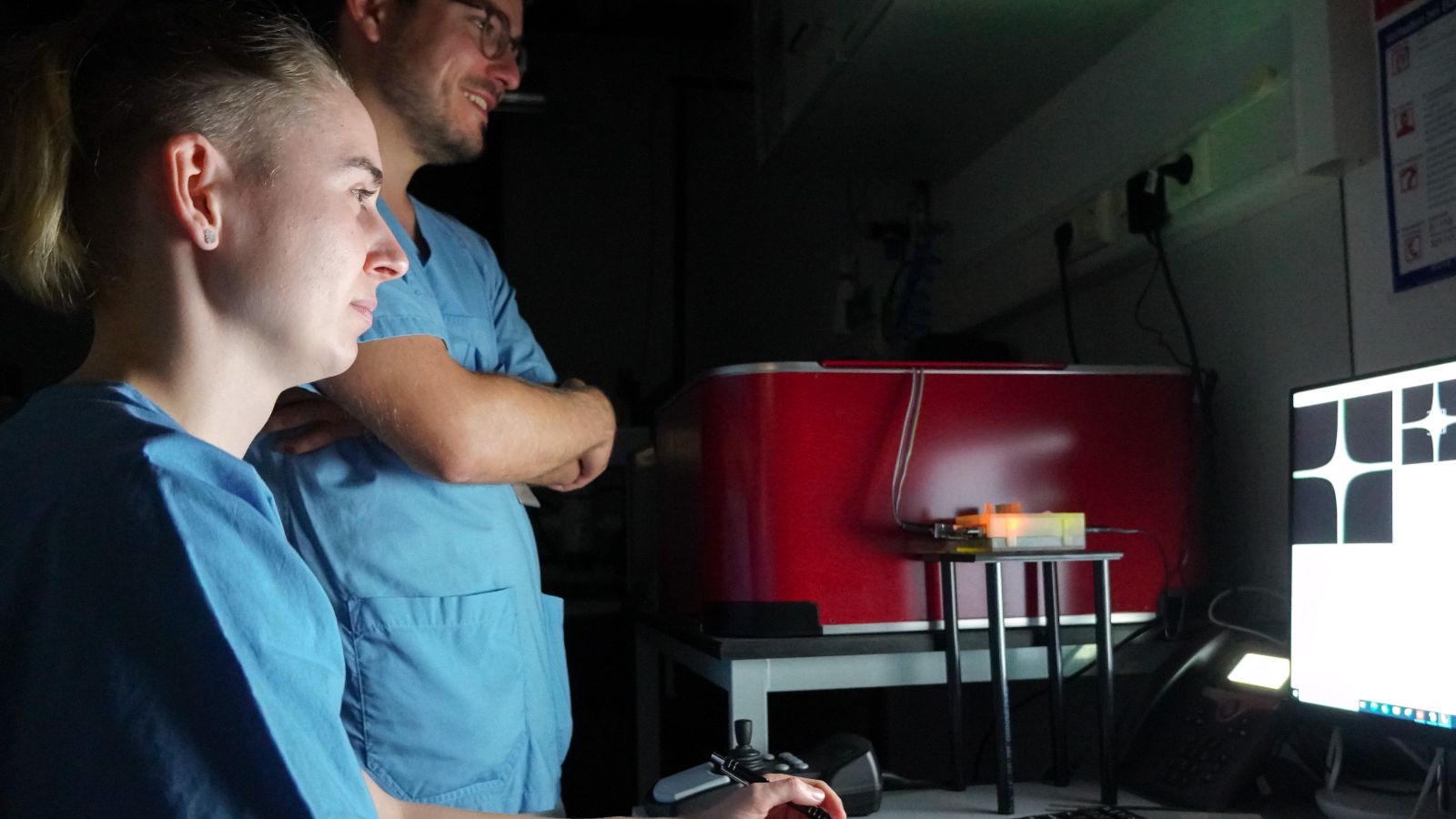 Zwei junge Forschende in blauen Laborkitteln, schauen auf eine Art Röntgenbild, das ein weißes Kreuz auf schwarzem Grund zeigt. Der Raum ist bis auf den Bildschirm mit dieser Darstellung dunkel.