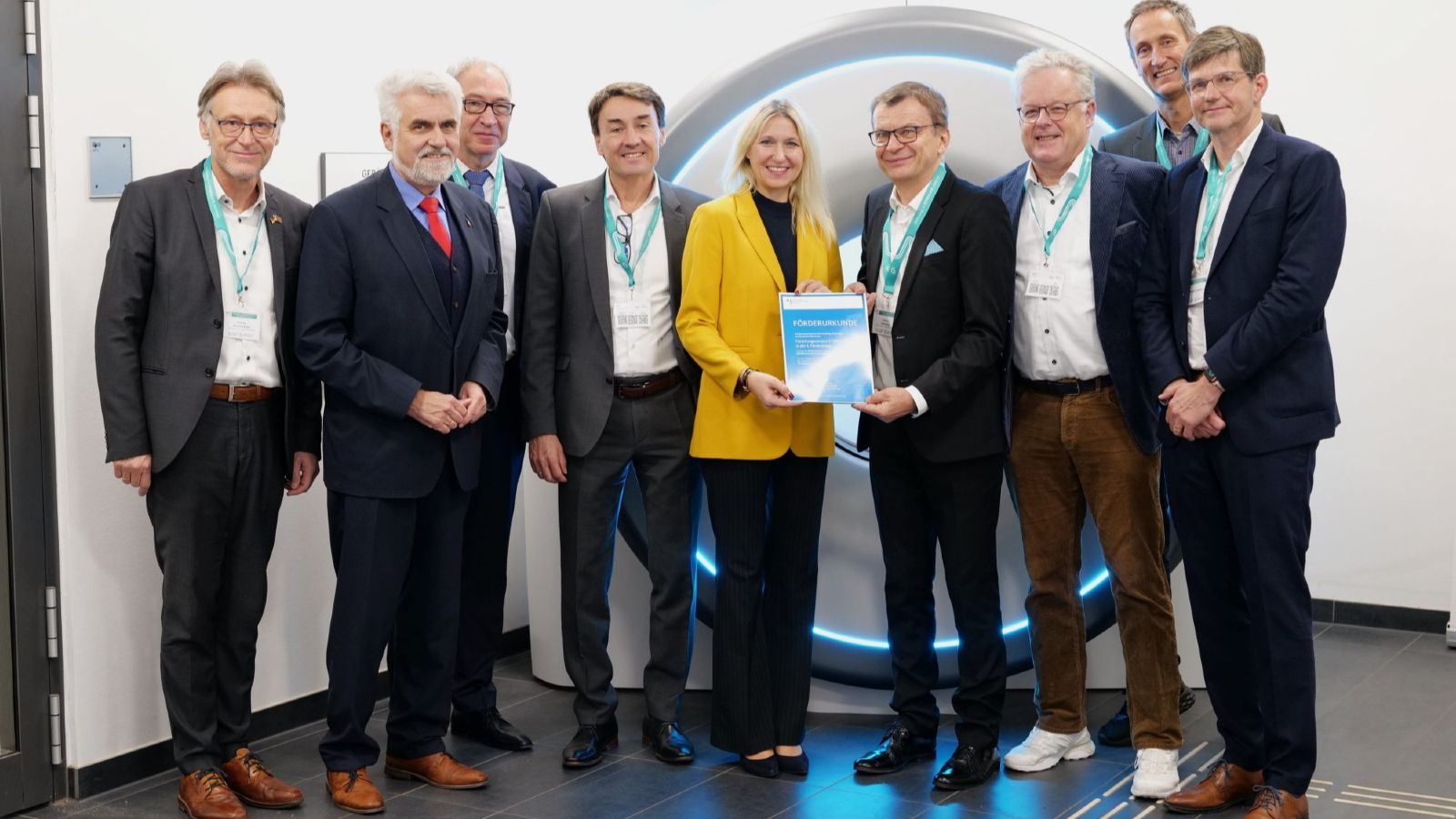 Die Parlamentarische Staatssekretärin Launert im gelben Jacket steht zwischen den Herren des STIMULATE-Vorstandes in schwarzen Jackets und überreicht eine Schmuckurkunde zum Übergang in die dritte Förderphase.