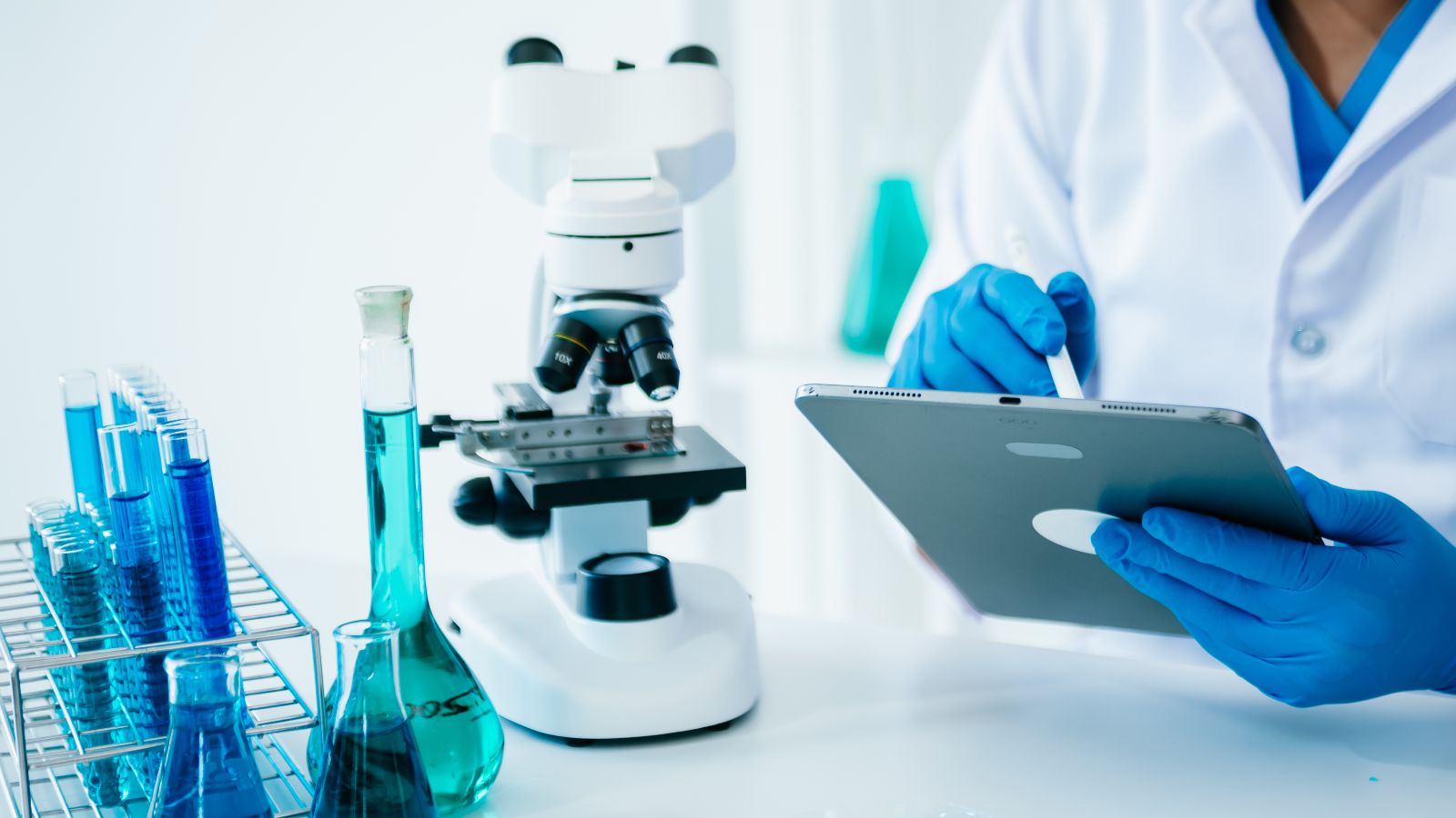 Flüssigkeiten, Mikroskop und etwas im Hintergrund ein Laborant mi Tablet